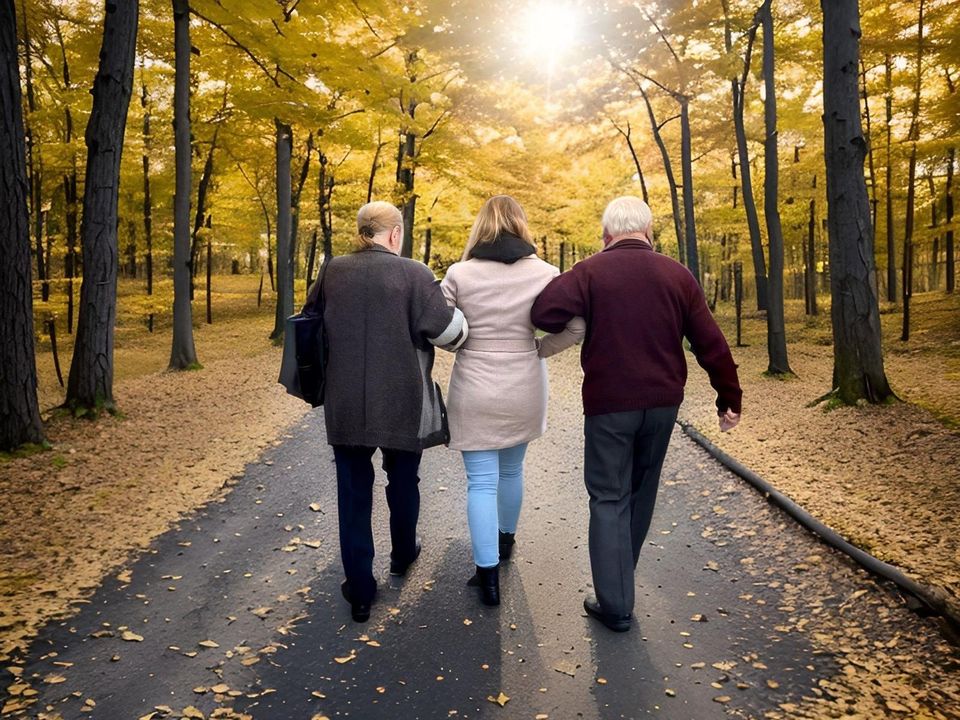 Eine Alltagsbegleiterin geht mit zwei älteren Menschen Arm in Arm durch einen herbstlichen Wald